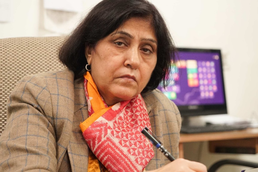 A woman with dark hair and wearing a coat with a red and white scarf holds a pen.