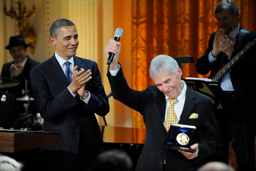 Barack Obama (left) applauds as Burt Bacharach (right) holds up mic with one hand and holds award with other.