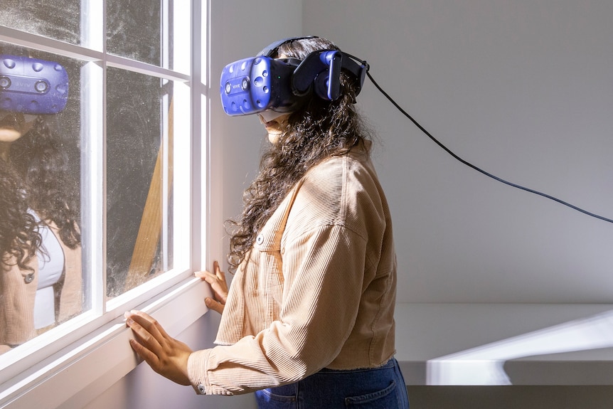 Woman with long dark curly hair wears taupe jacket, jeans and a blue virtual reality headset in white room in front of window.