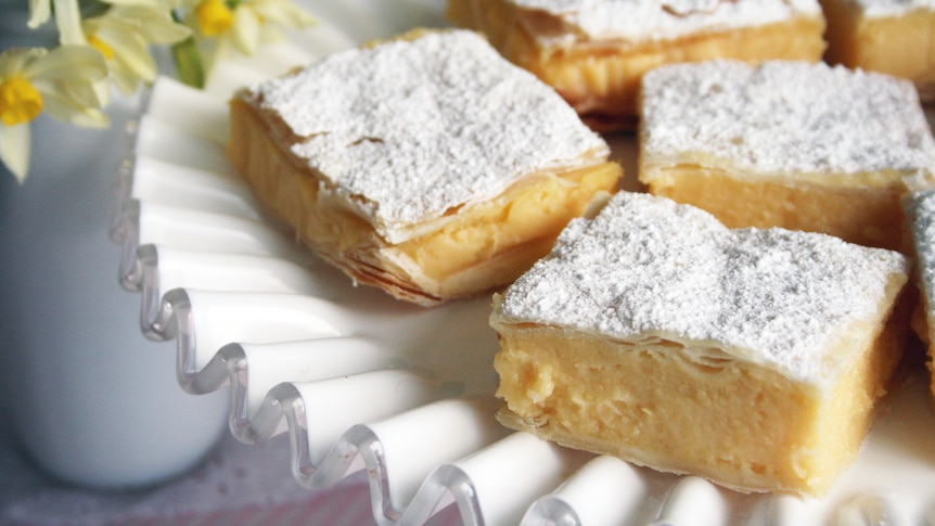 Vanilla slices sitting on a white decorative plate. The slices are dusted with icing sugar. There are flowers in the background.