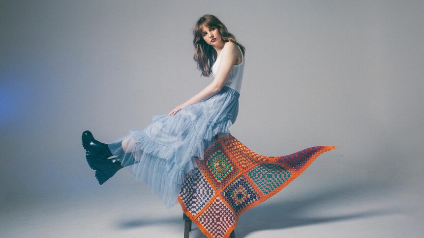 Gena Rose Bruce sits atop a stool covered by a knitted blanket, wearing a flowing skirt and boots.