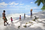 Three children walk along the beach with two adults, one carrying coconuts on her head.