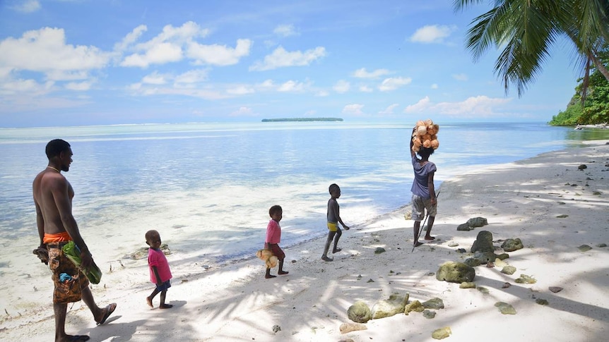 Three children walk along the beach with two adults, one carrying coconuts on her head.
