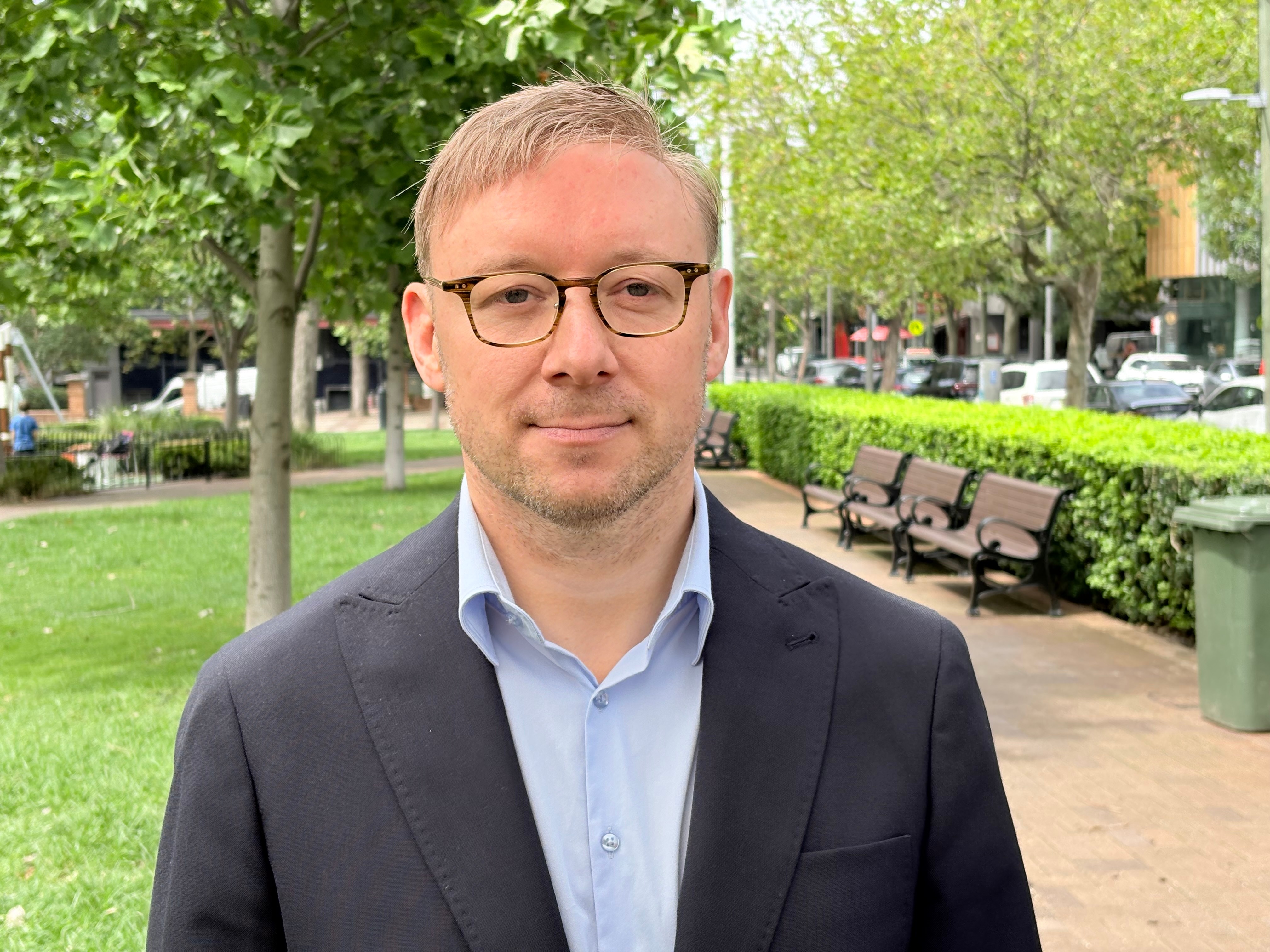 A person in a suit wearing glasses stands in a park smiling at the camera.