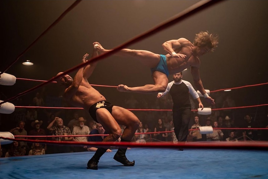 A film still of Zac Efron in the wrestling ring. He has 80s-style shaggy hair and is dropkicking his opponent.
