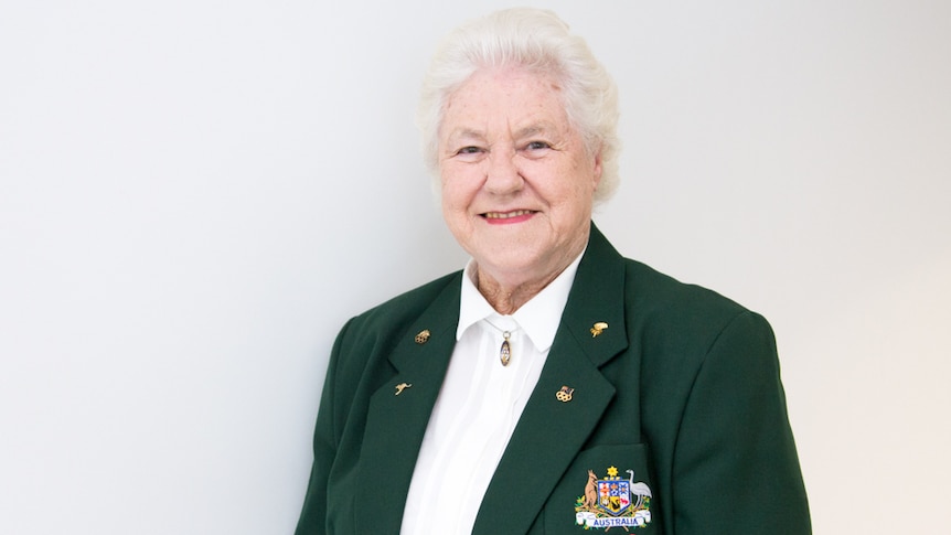 An older woman wears a green blazer embroidered with the words 'Olympic Games 1956'