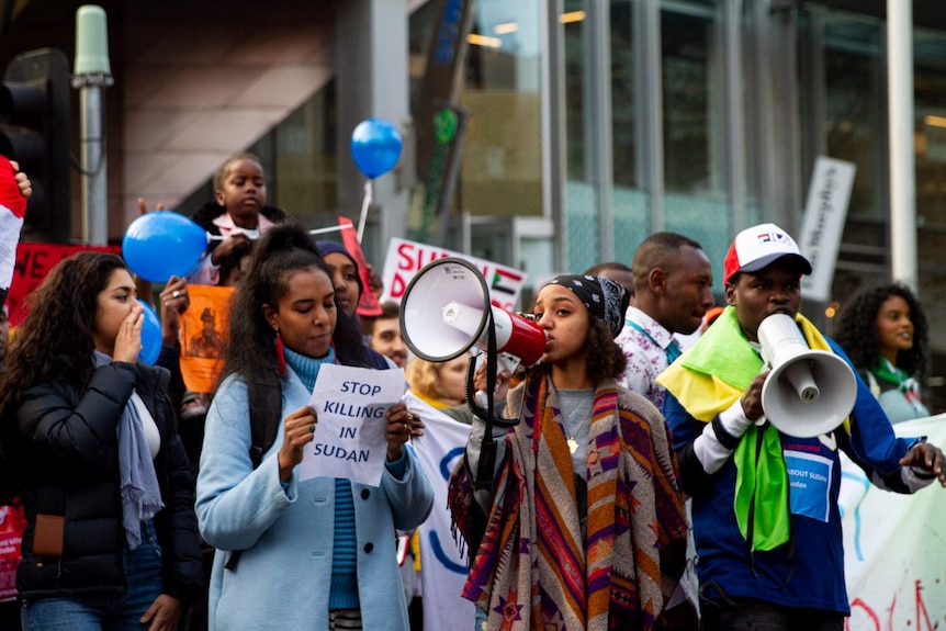 A large crowd of protesters holding megaphones, placards and blue balloons march down a street.