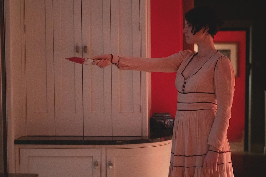 White woman with short dark hair wears long black trimmed peach dress and holds a bloody knife out in front of her.