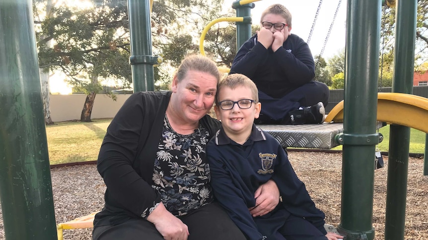 A woman hugs her son, who is wearing a school uniform. They are both sitting on a playground, with another child behind.