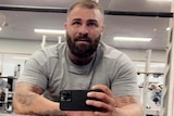 A muscular man with a thick beard and tattoos holds his mobile phone as he takes a mirror selfie at a gym