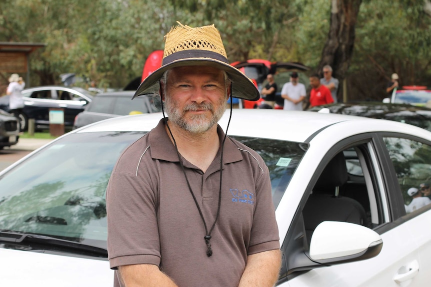A middle-aged white man in a polo short and broad-brimmed hat stands smiling against a white electric car.
