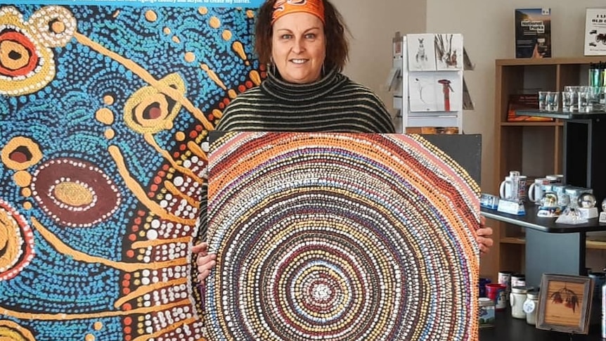 An indigenous artist stands with her painting in a gallery setting.