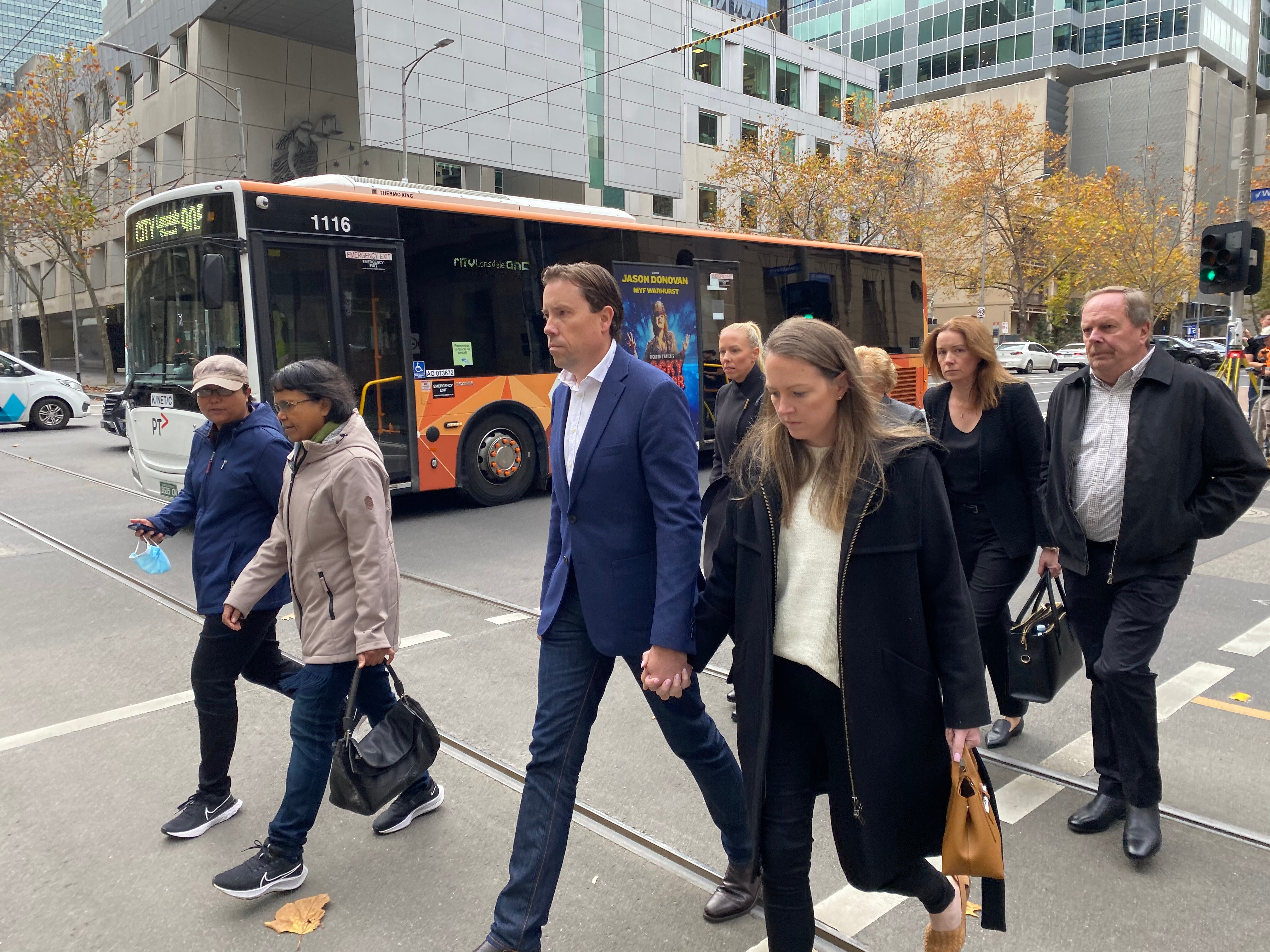 A group of people looking downcast crosses the road with a bus in the background