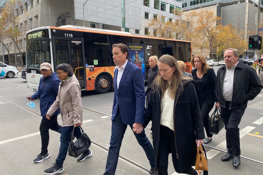 A group of people looking downcast crosses the road with a bus in the background