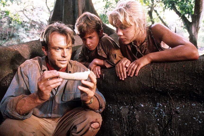 A still from the movie Jurassic Park showing Alan Grant holding a bone while two children look on, curious