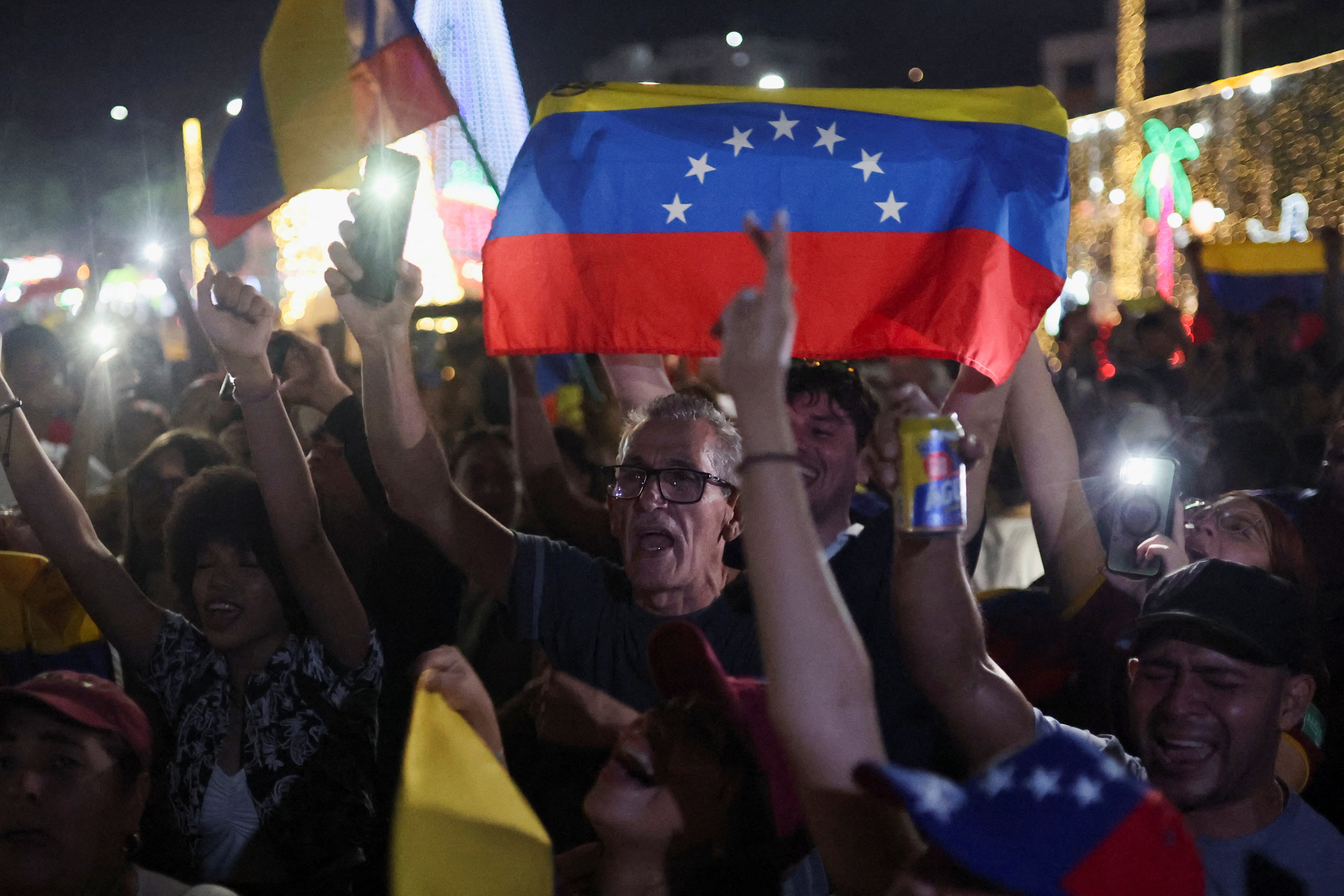 Un hombre sostiene un teléfono con una linterna y vitorea junto con una multitud que sostiene banderas rojas, azules y amarillas.