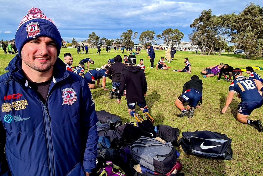 A man smiles to camera as rugby league players warm up in the background.