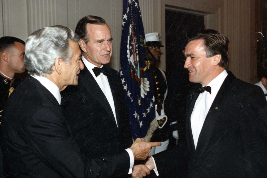 Hawke introducing Cassidy to Bush with all men in dinner suits and bow ties and US flag and soldier in background.