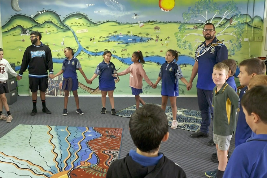 School children and teachers hold hands in a classroom.