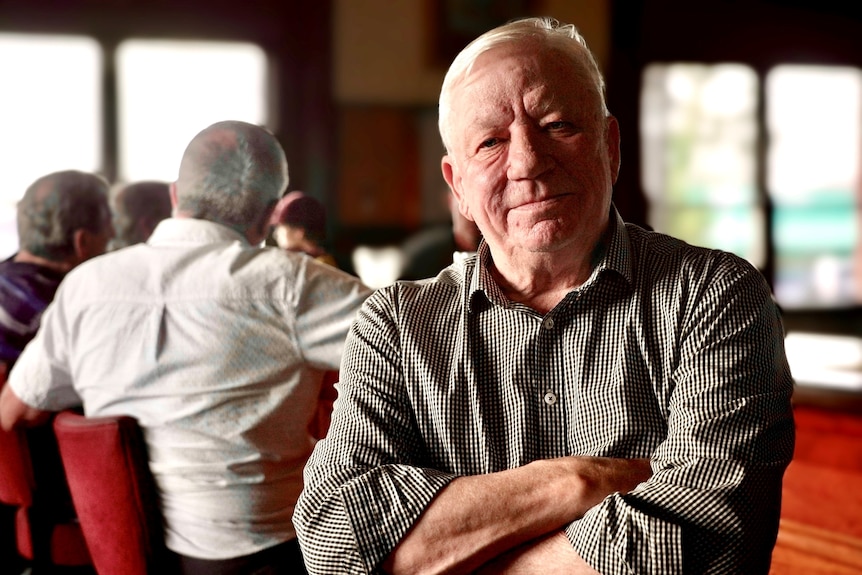 A grey-haired man in a grey, checked shirt stands, arms crossed, in a bar, with the backs of patrons visible behind him