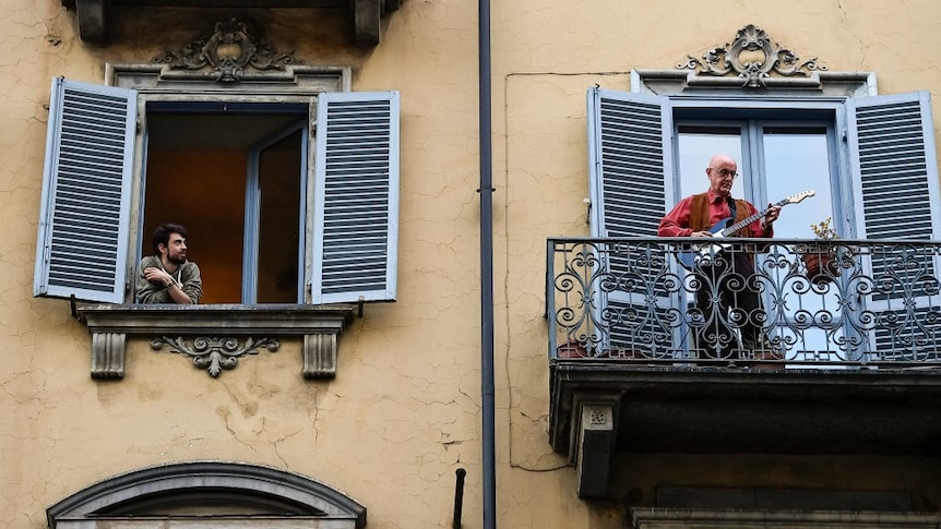 A man plays guitar on his balcony while a neighbour looks on