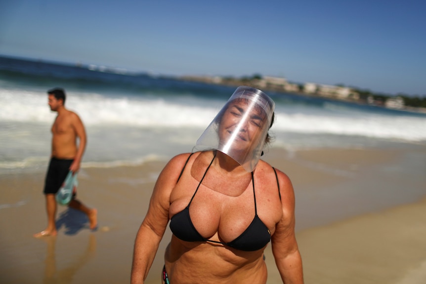 A woman wearing a black bathers and protective face shield walks along the Copacabana beach on a clear blue day.