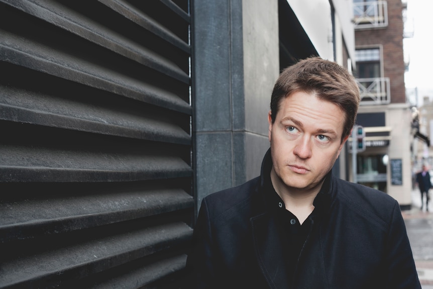 Johann Hari, with short, dark hair and black jacket, leans against an outside wall with slightly furrowed brow..