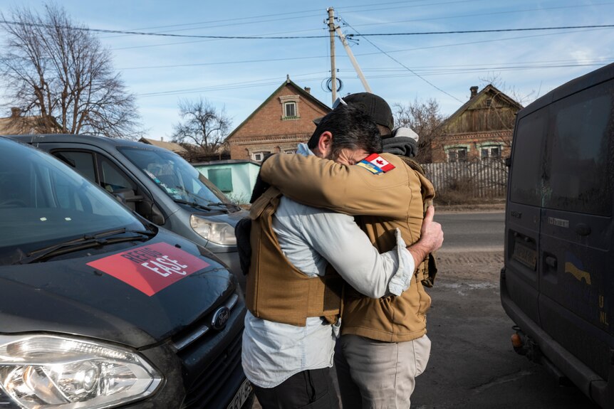 two men in body armour share a tight hug in front of vans