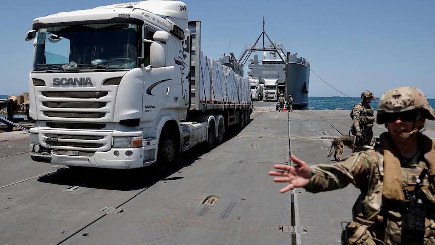 A soldier motions to the camera to move away as a huge truck drives into frame onto a pier off a huge boat