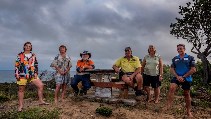 three women and three men sitting and standing around a bench with dark grey clouds in the background