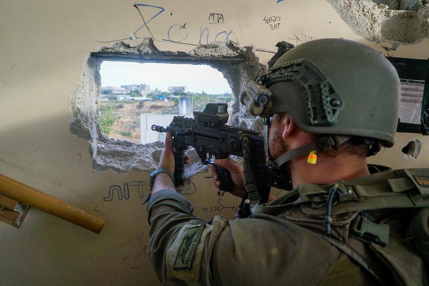 A close up of the back of an armedy Israeli soldier, wearing a helmet and pointing a gun through a square hole in a wall