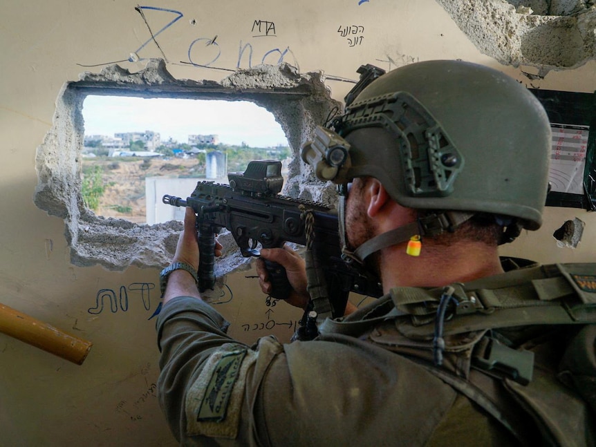 A close up of the back of an armedy Israeli soldier, wearing a helmet and pointing a gun through a square hole in a wall