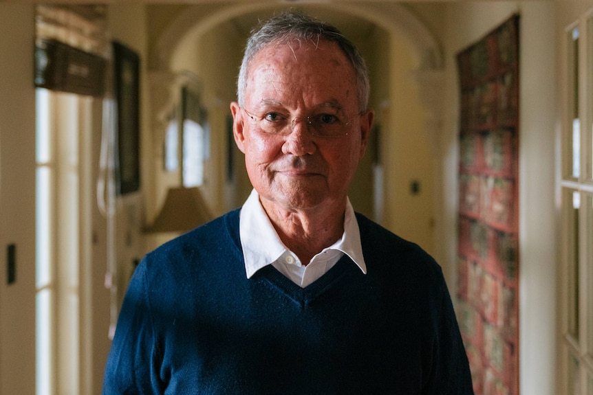 An older man wearing glasses stands in front of the camera.