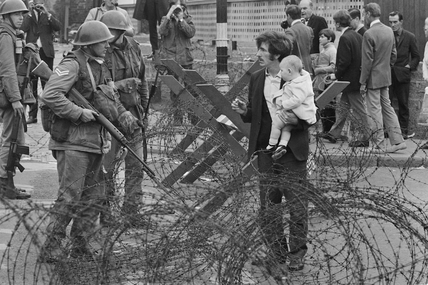 A 1960s black and white photo of a father and baby standing at a military road stop with soldiers and barbed wire