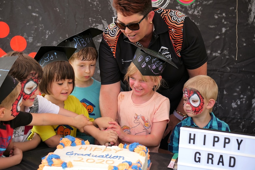 A woman cutting big sponge cake that says 'HIPPY graduation 2020' with six kids around her.