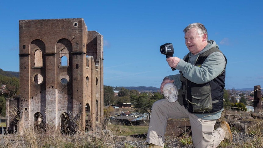 A man holding a skull and looking through an infrared viewer kneeling in front of an old ruined industrial building