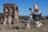 A man holding a skull and looking through an infrared viewer kneeling in front of an old ruined industrial building