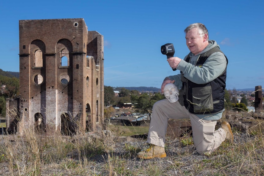 A man holding a skull and looking through an infrared viewer kneeling in front of an old ruined industrial building