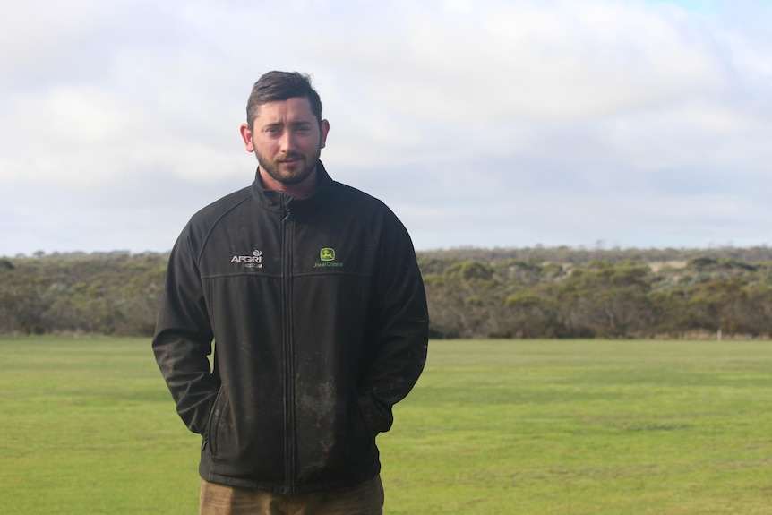 A bearded, dark-haired man wearing a dark jacket with two logos, stands in a grassy paddock with a line of trees in background