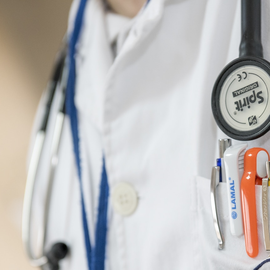 A doctor wearing a white lab coat with pens in their pocket and a stethoscope. Chest only in frame