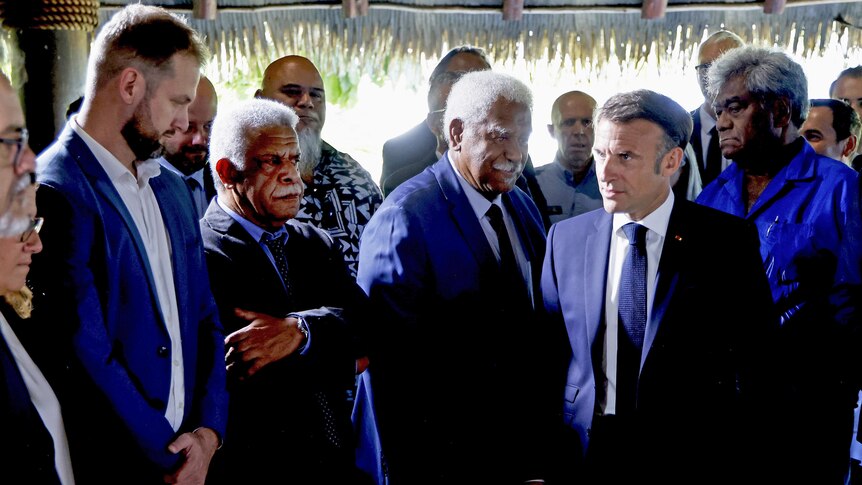 A group of men wearing suits, standing up, greeting the French president. One man has his arms crossed.