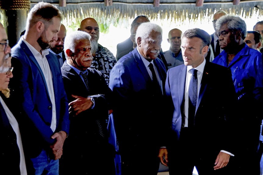 A group of men wearing suits, standing up, greeting the French president. One man has his arms crossed.