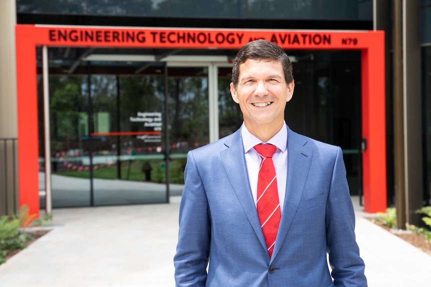 A man in a suit and tie stands in front of a university building, smiling.