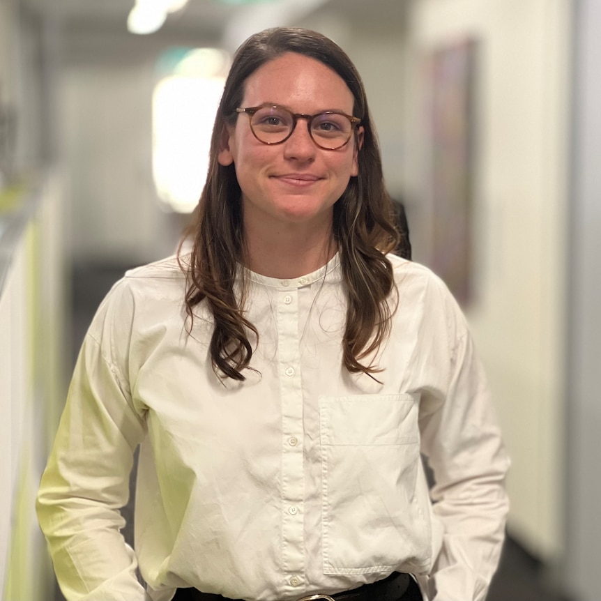 A woman in a white shirt with glasses looks directly into the camera.