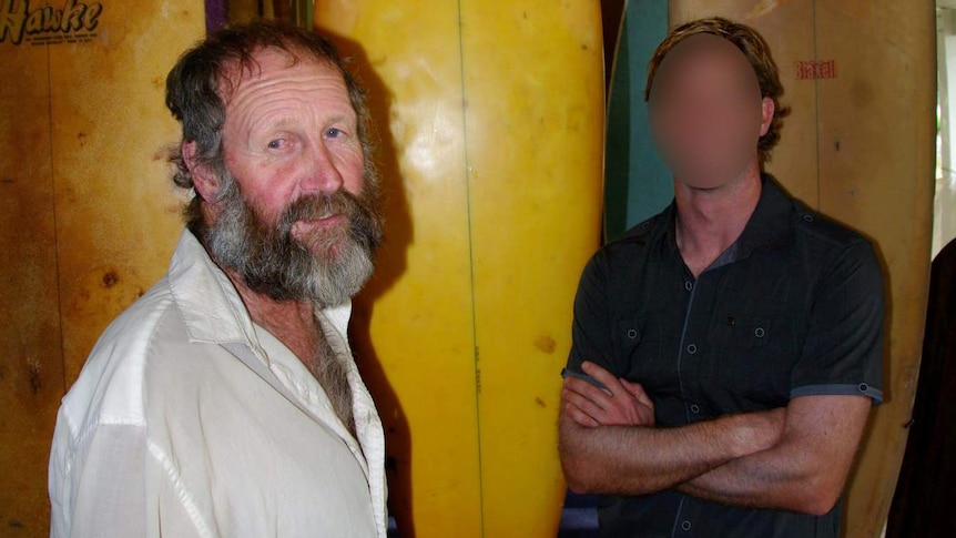 Antony Hardy pictured in front of surf boards. He was a surfboard maker. Another man stands near him, his face is blurred.