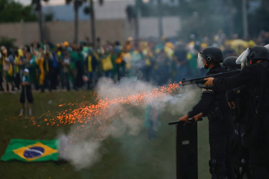 Security operatives wearing full riot gear fire their weapons, a crowd and a crumpled Brazilian flag on the ground behind