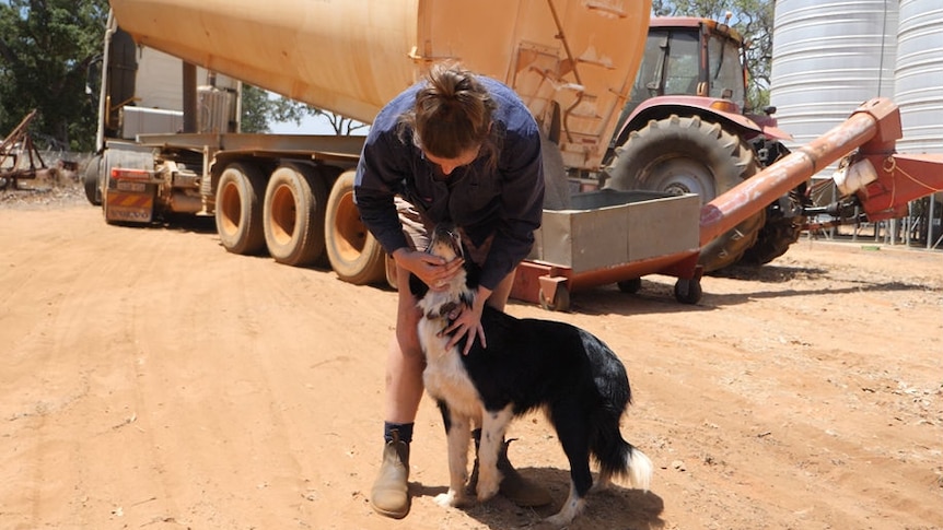 A woman bends over and pats her dog as they stand together on a dusty road in front of a parked truck.