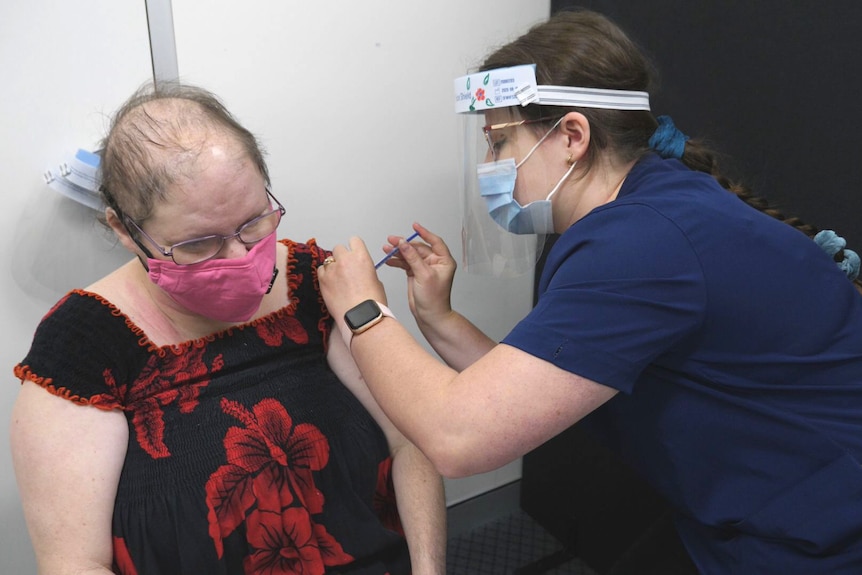Raelene wearing a pink face mask as a nurse injects her arm with a vaccine.