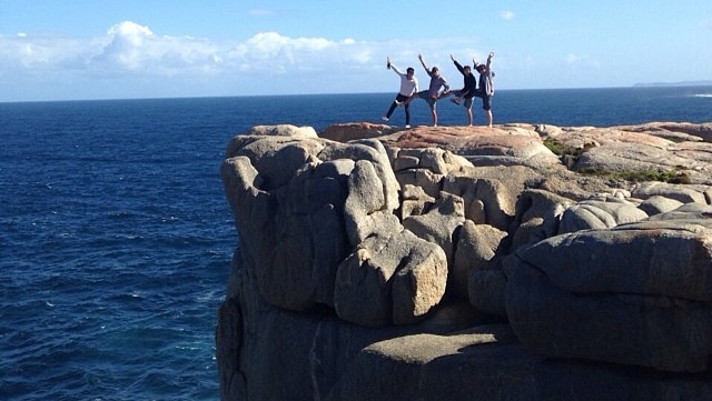 A photo of a group of four people standing on the edge of a cliff face posing with one leg and two arms up in the air.