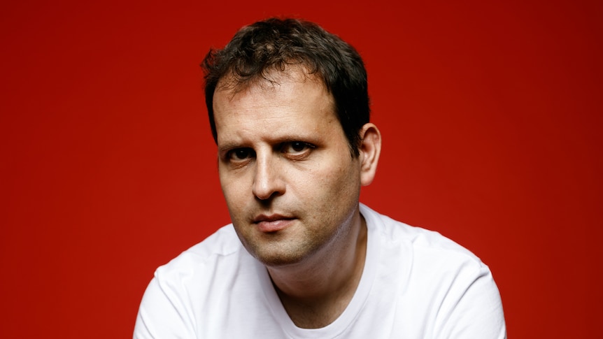 A portrait of Adam Kay, a 43-year-old man, leaning forward while perched on a stool in front of a red background.